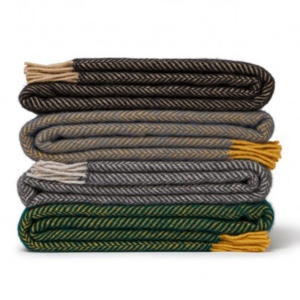A stack of five folded woven throw blankets with herringbone patterns in black, gray, green, and brown, each with contrasting tassels, set against a plain white background.