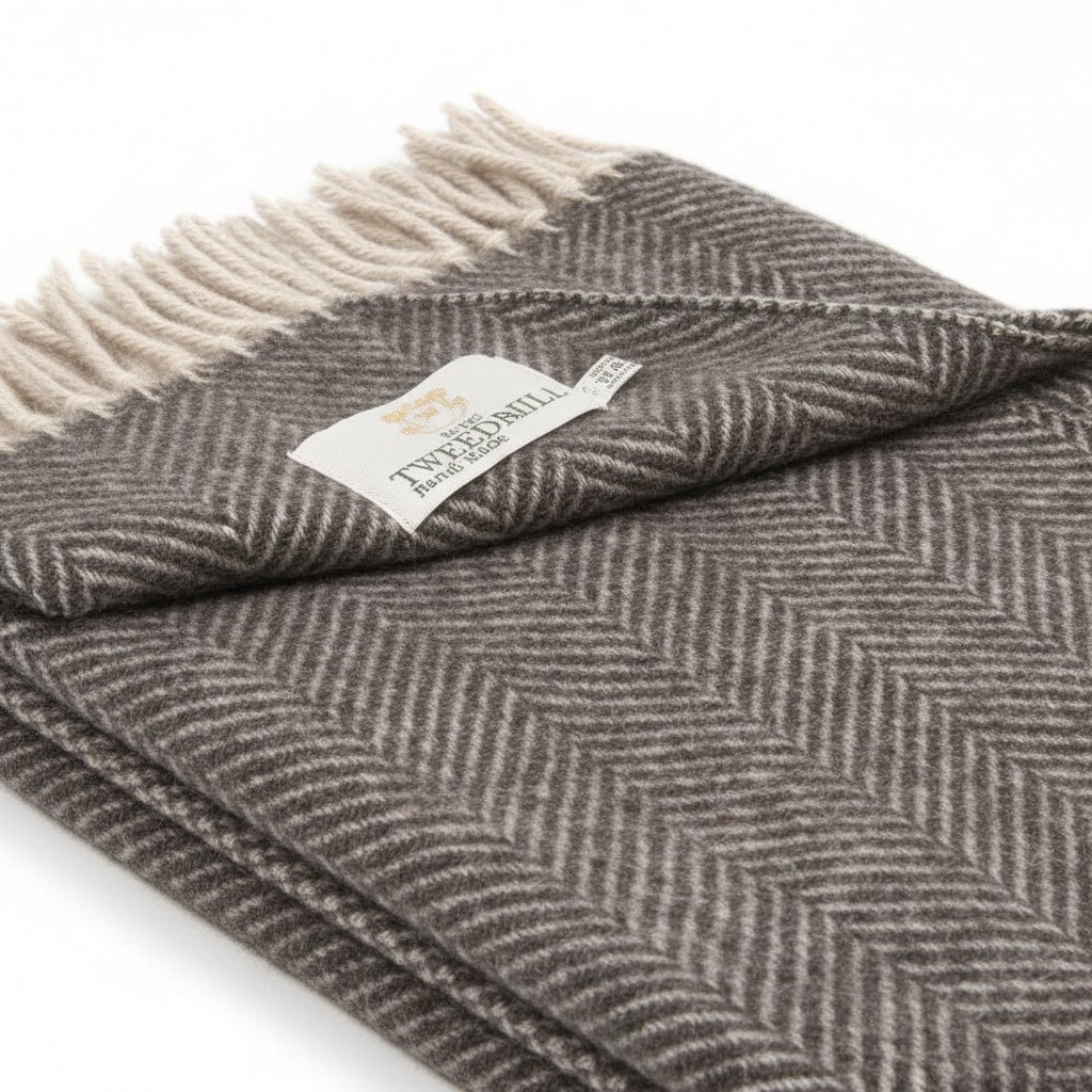 A folded gray and beige herringbone woven throw blanket with cream fringe and a small white brand tag, shown on a plain white background.