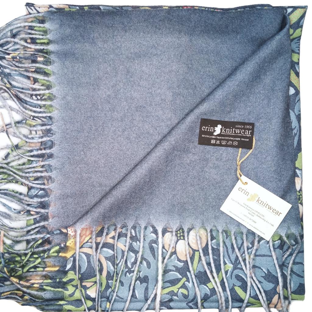 A folded knit scarf in a muted slate blue-gray color, featuring a detailed botanical border pattern in green, blue, and beige tones. The scarf has fringe on one side and a black brand label reading 'erin knittwear' attached with a string.