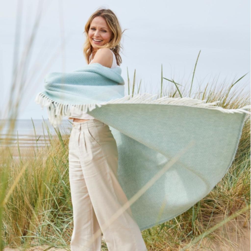 Woman on a beach wearing a light blue Tweedmill Beehive Ocean wool throw with fringed edges.